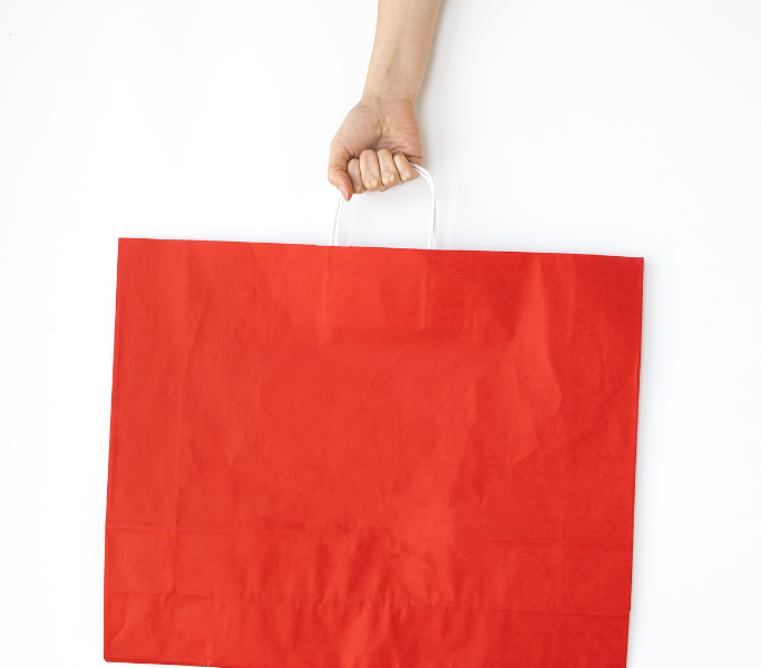 Sacola de compras de papel vermelho, em formato retangular, com alça centralizada na abertura da sacola, sendo uma mão que a segura pela alça.